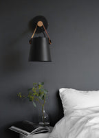 Black Clérachic wall light on a charcoal wall above a bed with white linen, a glass vase of leaves and a magazine.
