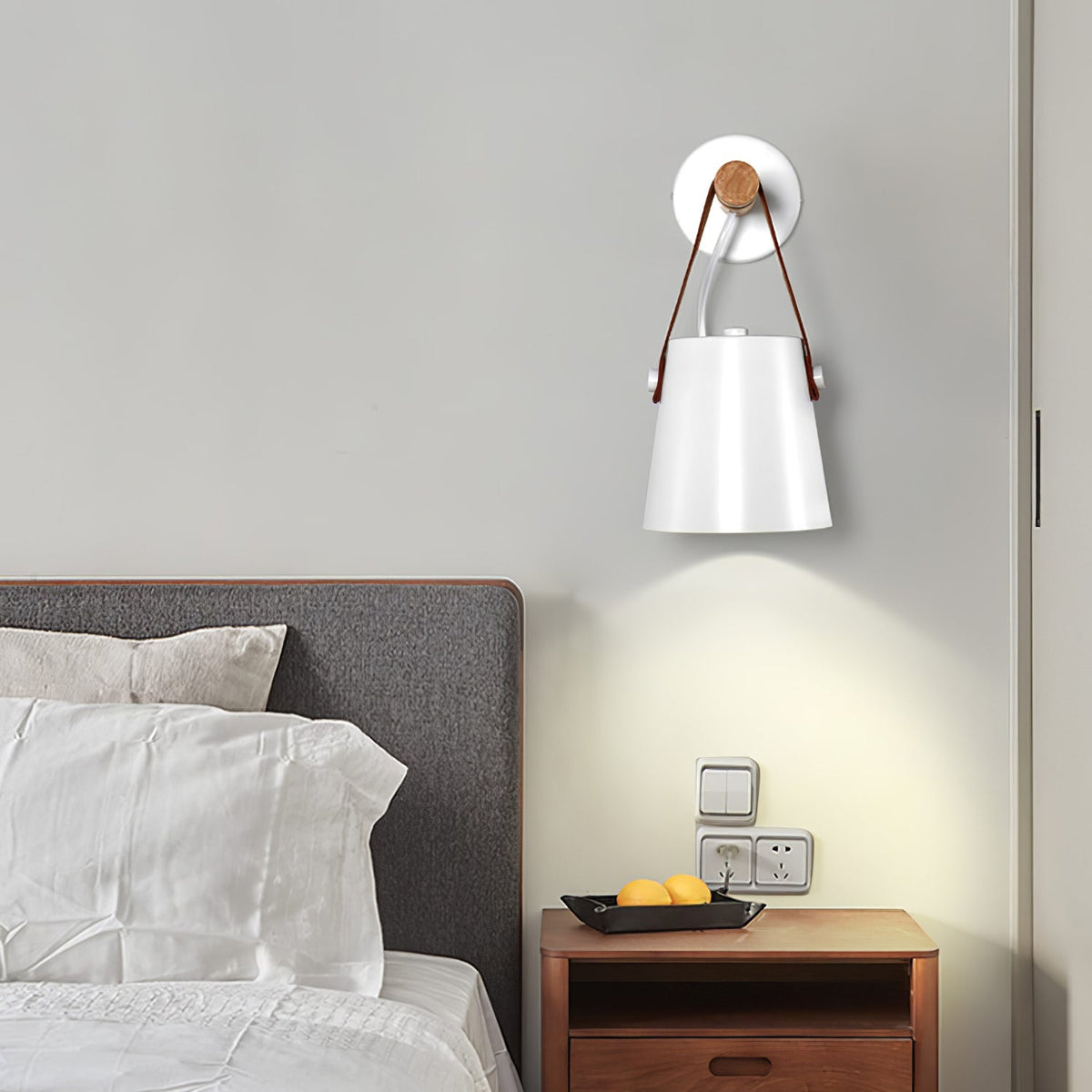 White Clérachic wall light above a bedside table with a bowl of lemons, beside a bed with white pillows and a grey headboard.