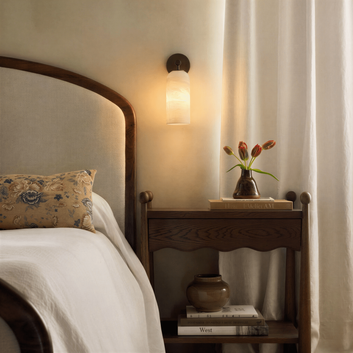 Clérachic wall lamp in bronze finish lit beside a bed, above a wooden nightstand holding tulips in a stoneware vase.