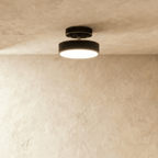 Clérachic matte black flush-mount ceiling light with a warm frosted disc, shown centred on a plain plaster ceiling.
