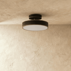 Clérachic matte black flush-mount light fixture glowing warm, set against a textured beige plaster ceiling and wall.