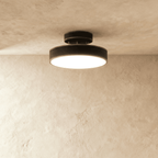 Clérachic black flush-mount light with a rounded drum shade, glowing warm beneath a beige plaster ceiling corner.