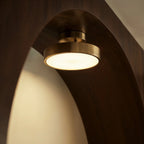 Clérachic copper flush-mount ceiling light shown close beneath a curved wood archway, its disc glowing warm white.