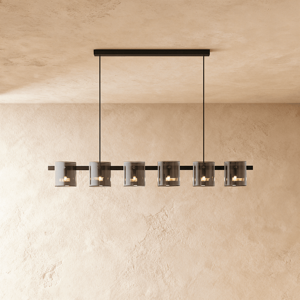 Clérachic linear chandelier with six smoky glass cylinder shades along a slim black bar, against a plaster wall.