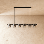 Clérachic linear chandelier with six smoky glass cylinder shades along a slim black bar, against a plaster wall.