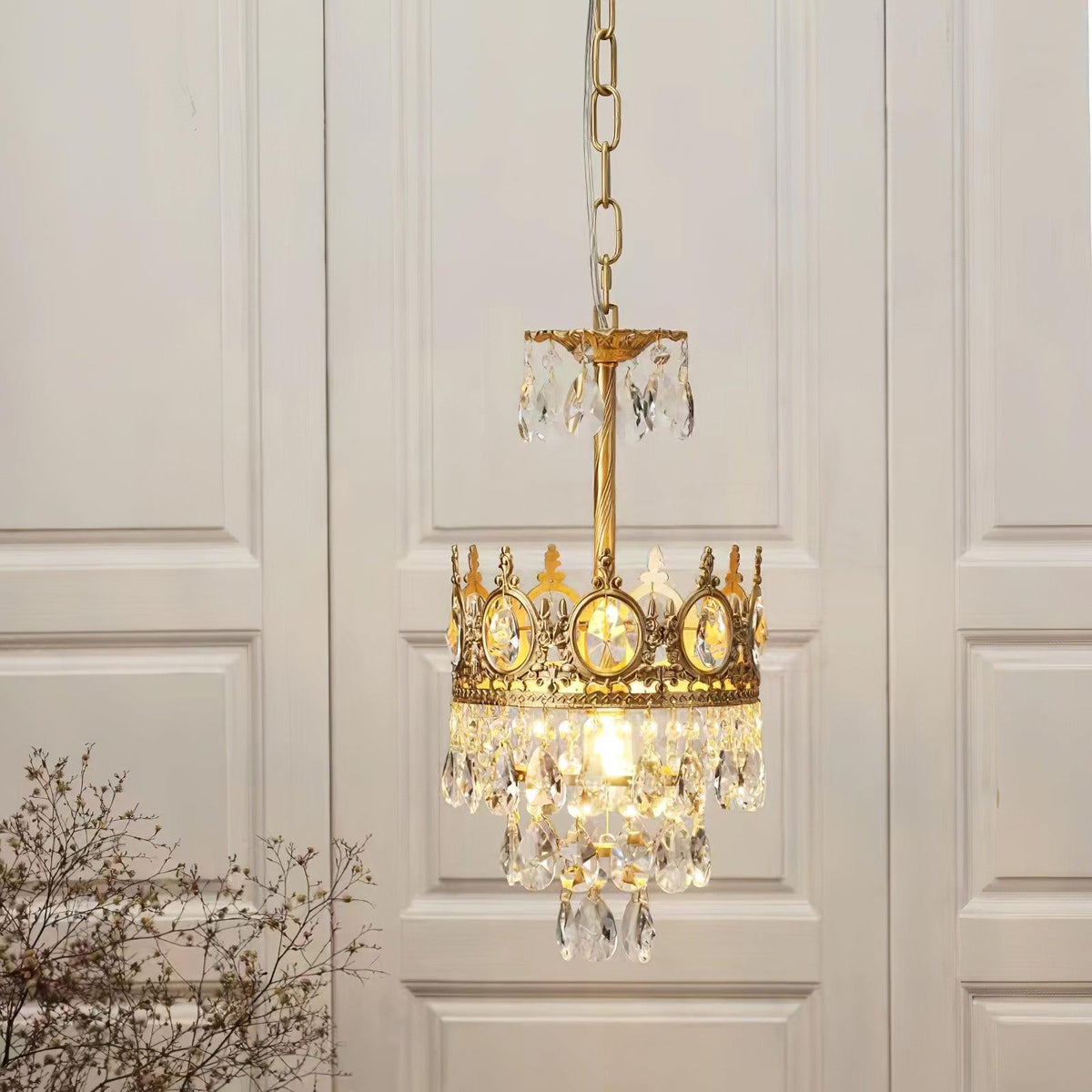 Smaller single-tier Clérachic crystal chandelier hung on a chain in front of white panelled doors, with a bare branch at left.