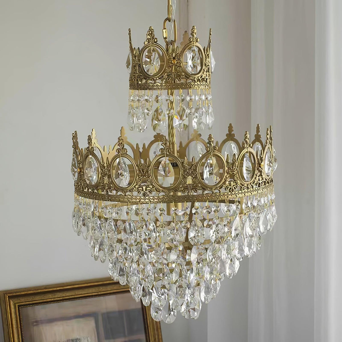Clérachic crystal chandelier beside a sheer curtain and a gilt picture frame, with crown-shaped tiers and clear drops.