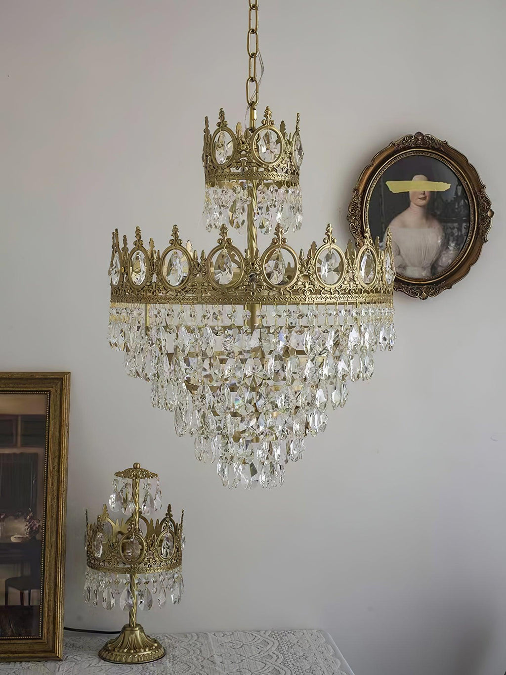 Clérachic crystal chandelier on a white wall beside an oval portrait, with a small crown-shaped table lamp below.