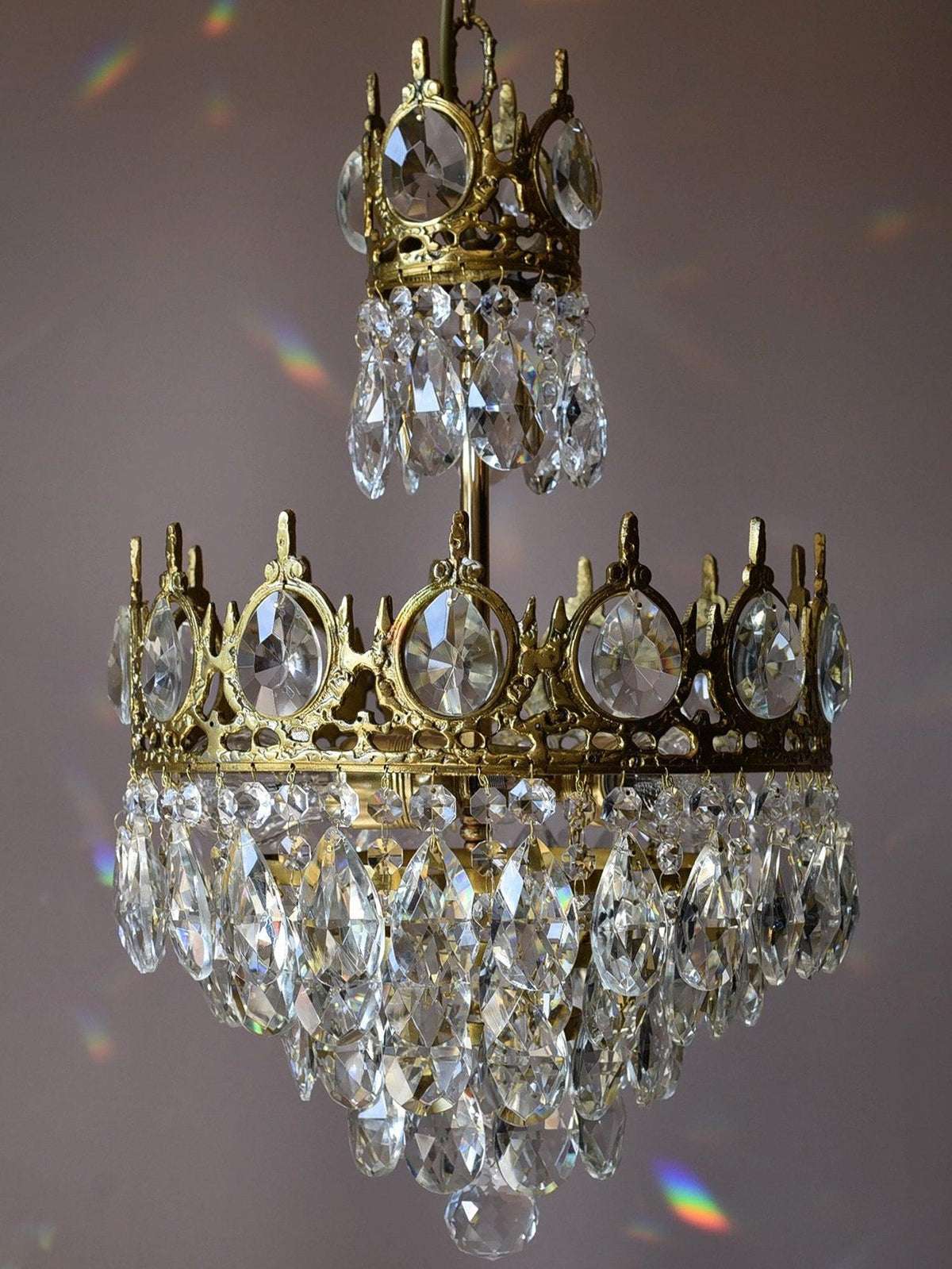Unlit Clérachic chandelier against a mauve backdrop, showing its crown-edged tier and the clear teardrop crystals.