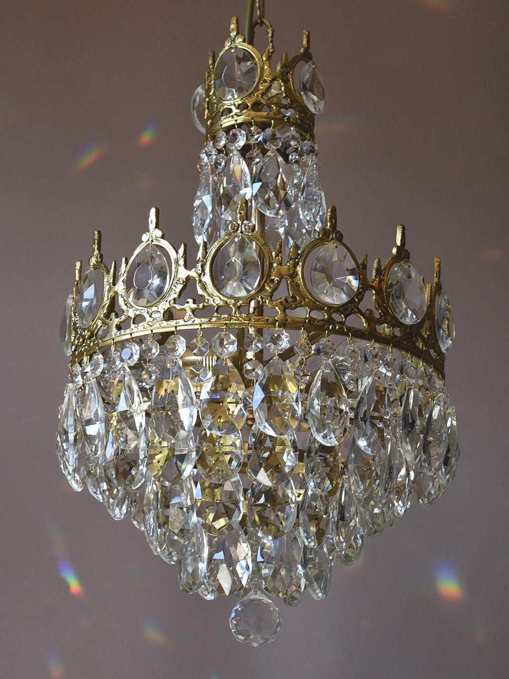 Side view of this Clérachic chandelier on a mauve backdrop, with a small crown tier above a larger tier of crystals.
