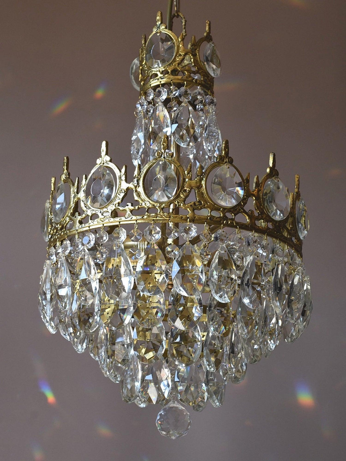 Side view of this Clérachic chandelier on a mauve backdrop, with a small crown tier above a larger tier of crystals.