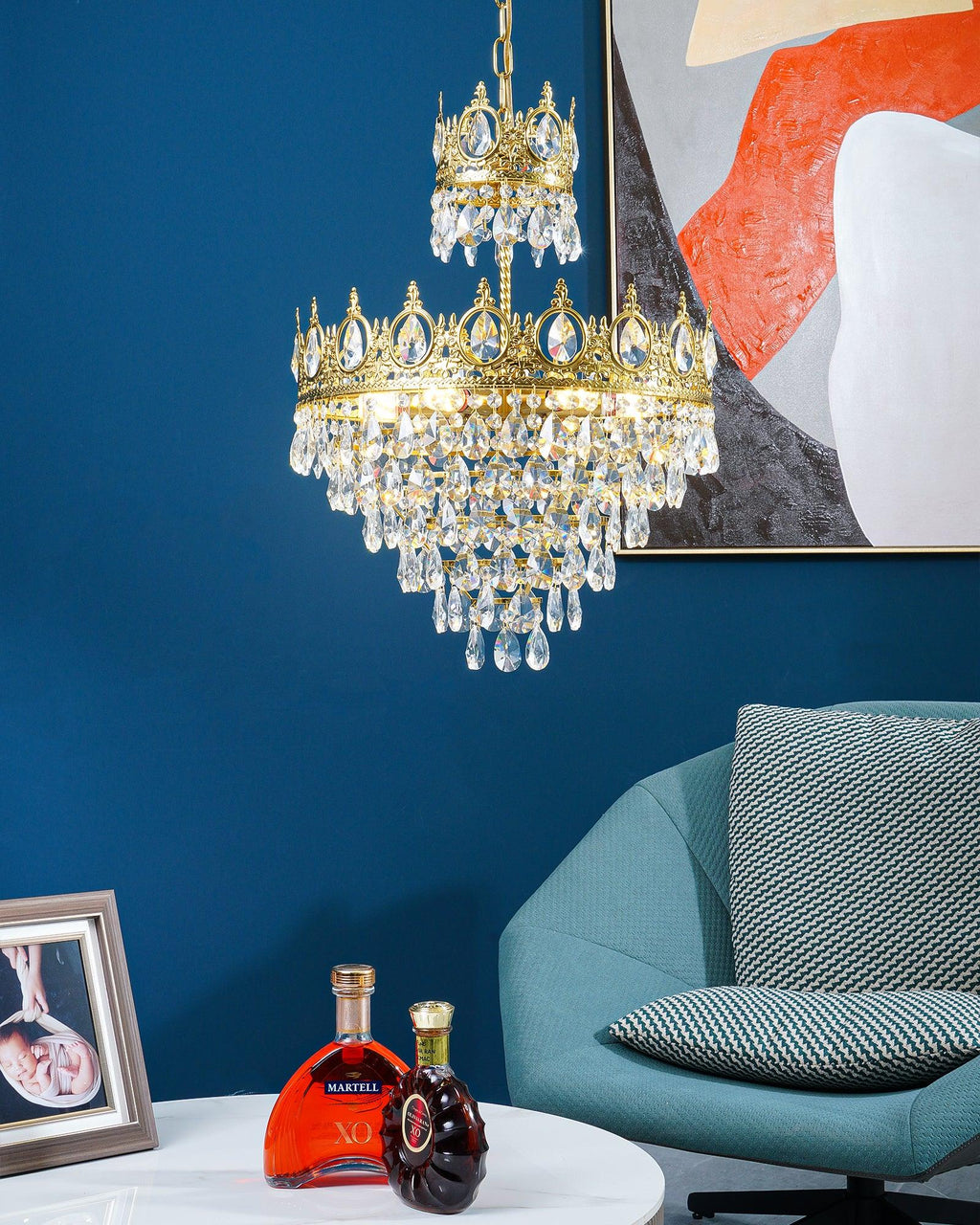 Clérachic crystal chandelier against a deep blue wall and abstract print, above a white table with two bottles and a teal chair.