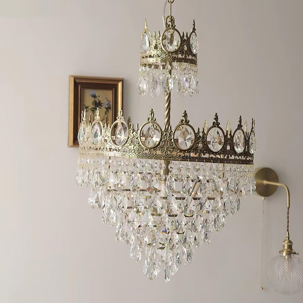 Clérachic crystal chandelier on a pale wall beside a small framed picture and a brass wall light with a glass shade.