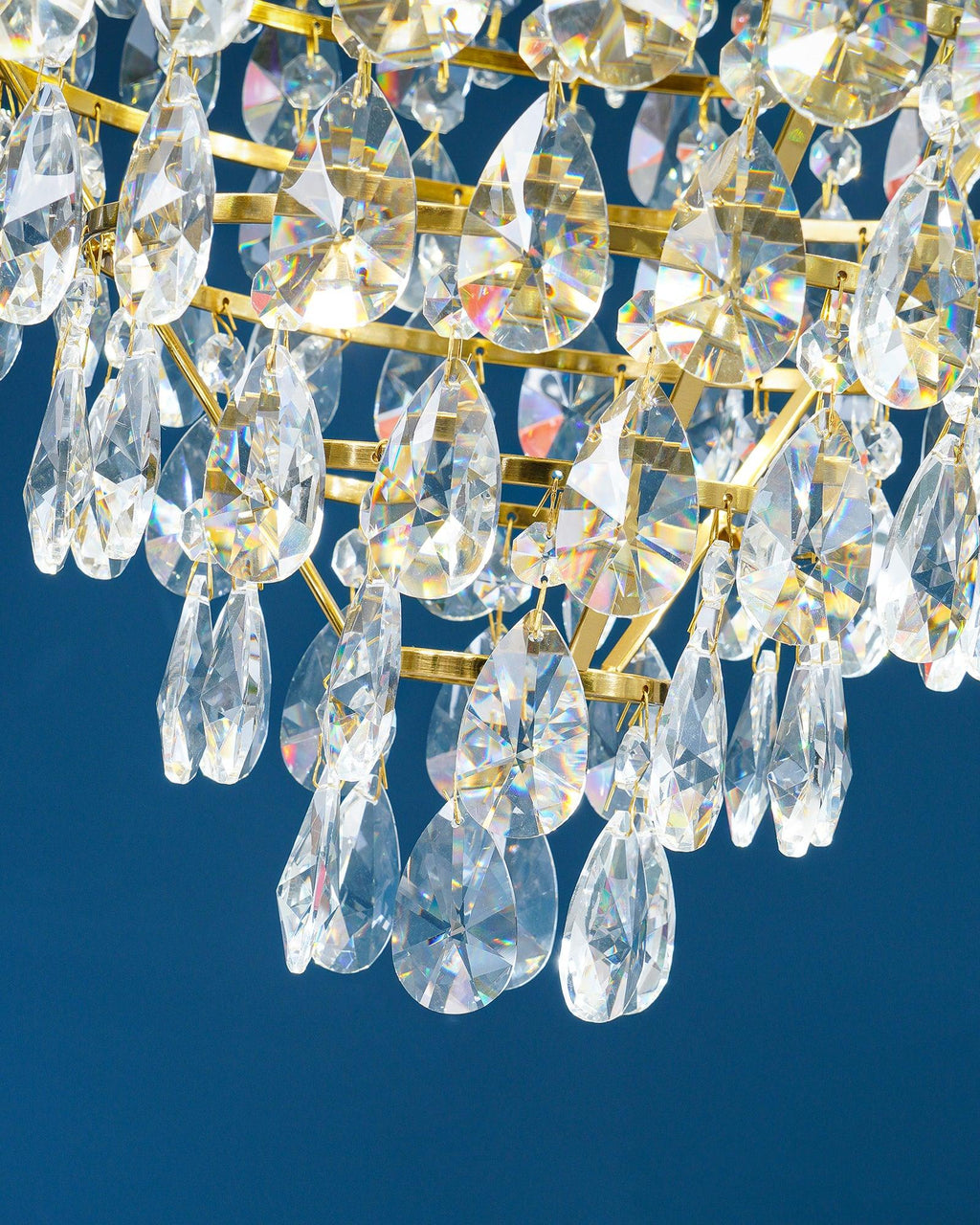 Close view of layered clear crystal drops and brass rings under the crown tier of this Clérachic chandelier, on blue.