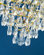 Close view of layered clear crystal drops and brass rings under the crown tier of this Clérachic chandelier, on blue.