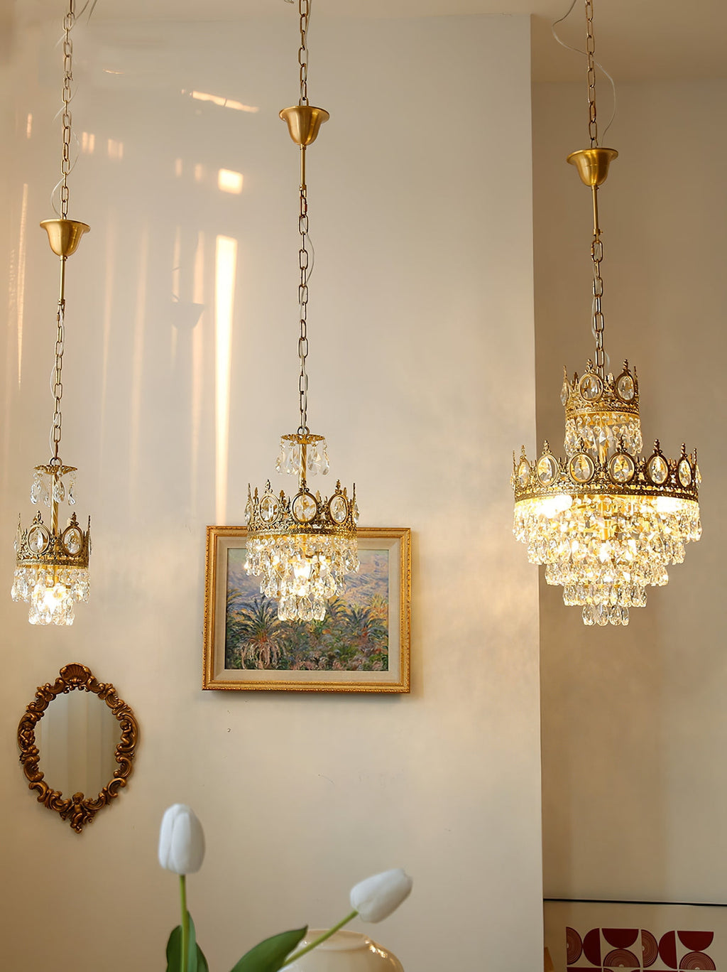 Three Clérachic chandeliers of different sizes hanging on chains in a pale hall, with a framed painting and tulips below.