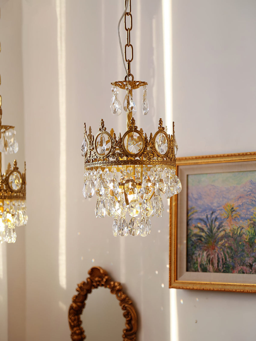 Smaller Clérachic chandelier on a chain, beside a gilt-framed painting of palms and a small ornate mirror.