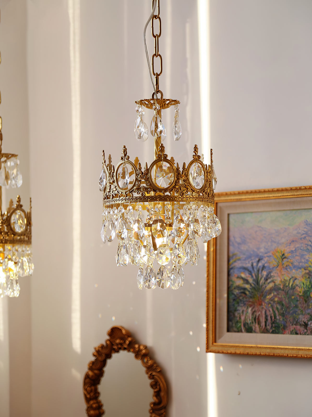Smaller Clérachic chandelier on a chain, beside a gilt-framed painting of palms and a small ornate mirror.