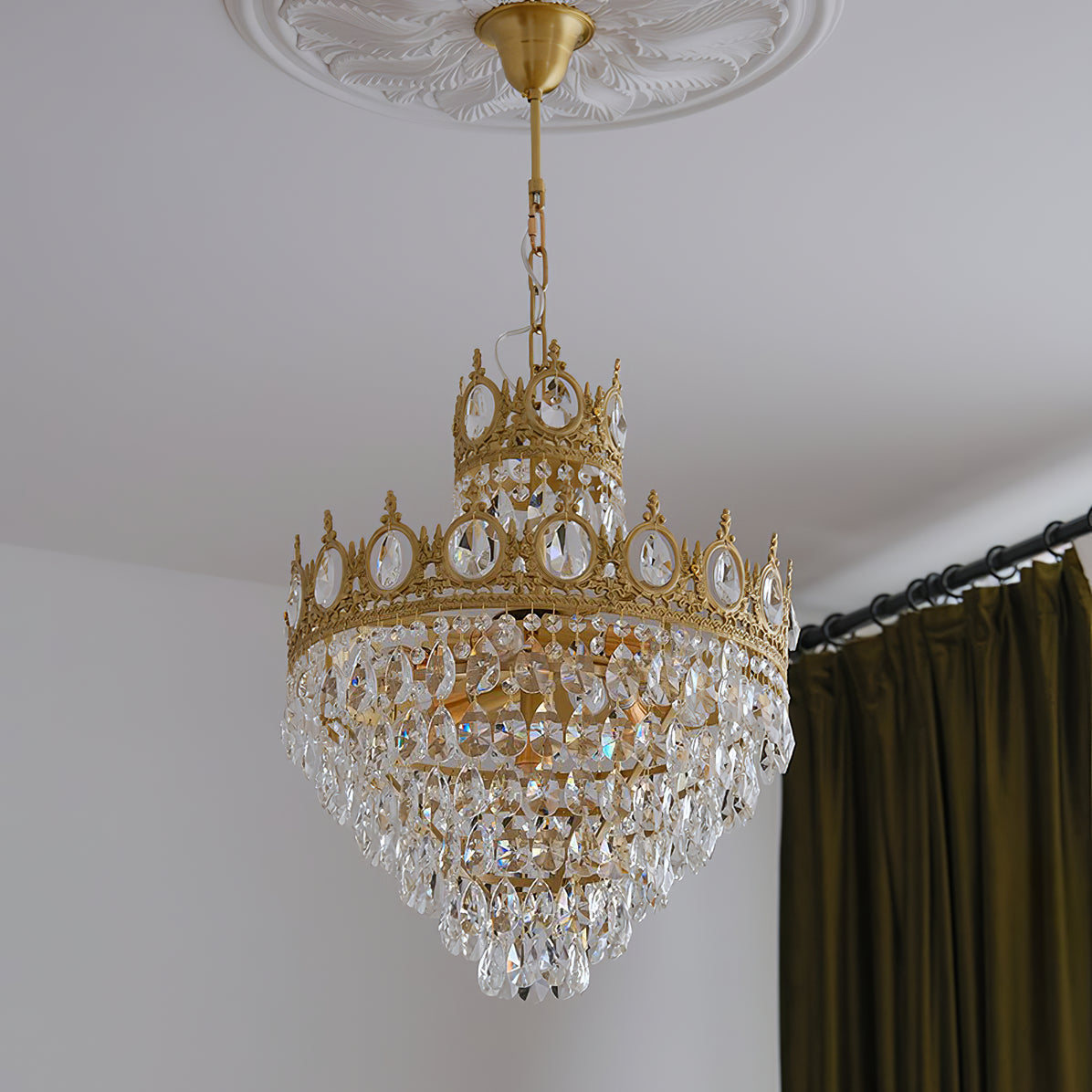 Clérachic crystal chandelier hung from a brass canopy beneath a plaster ceiling rose, with an olive-green curtain beside it.
