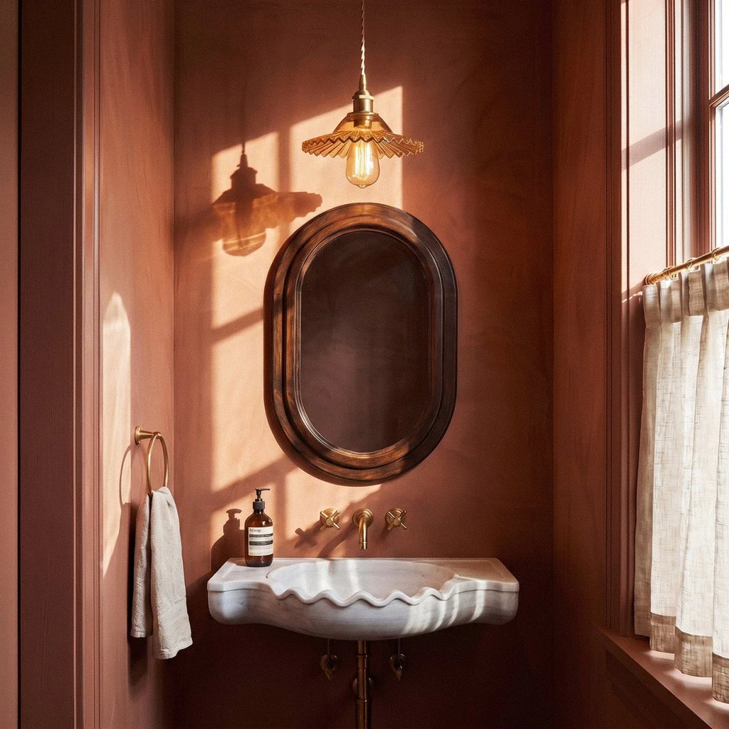 A Clérachic Isabel pendant light hangs above a marble sink in a warm terracotta powder room beside an oval mirror.