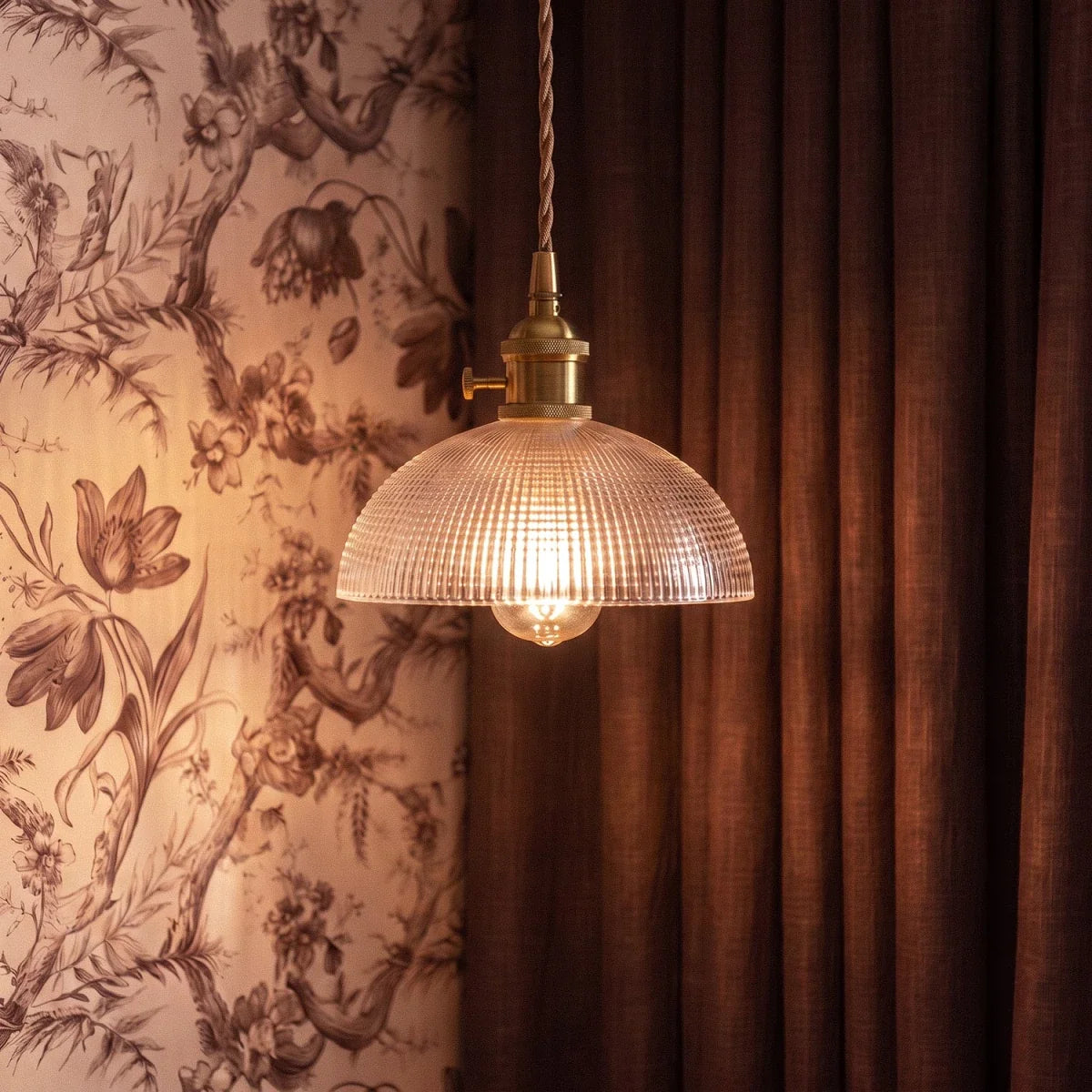 A Clérachic Isabel pendant light with a woven grid glass shade hangs against dark curtains and floral toile wallpaper.