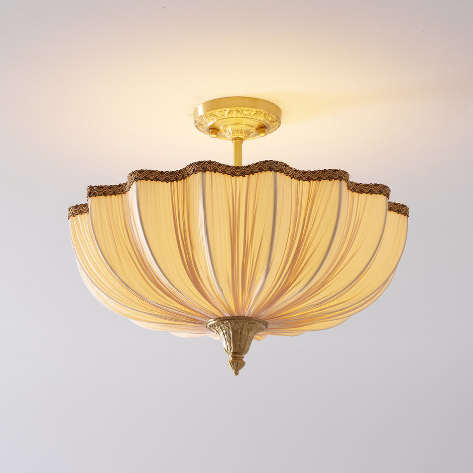 Front view of the lit Clérachic ceiling light, its pleated cream fabric glowing warm beneath a brass ceiling plate.