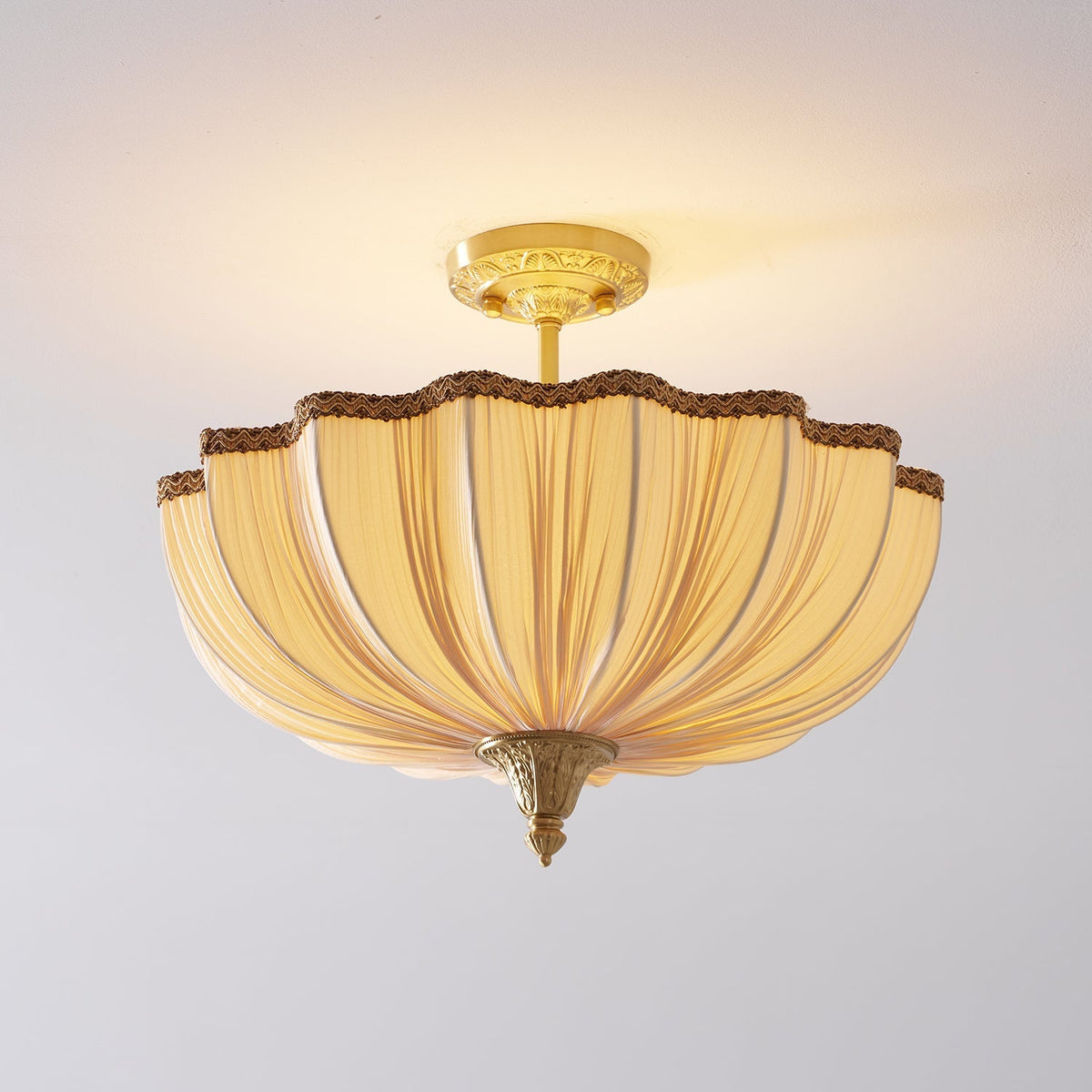 The Clérachic ceiling light lit, a scalloped fabric edge with a braided trim and a brass finial at the base.