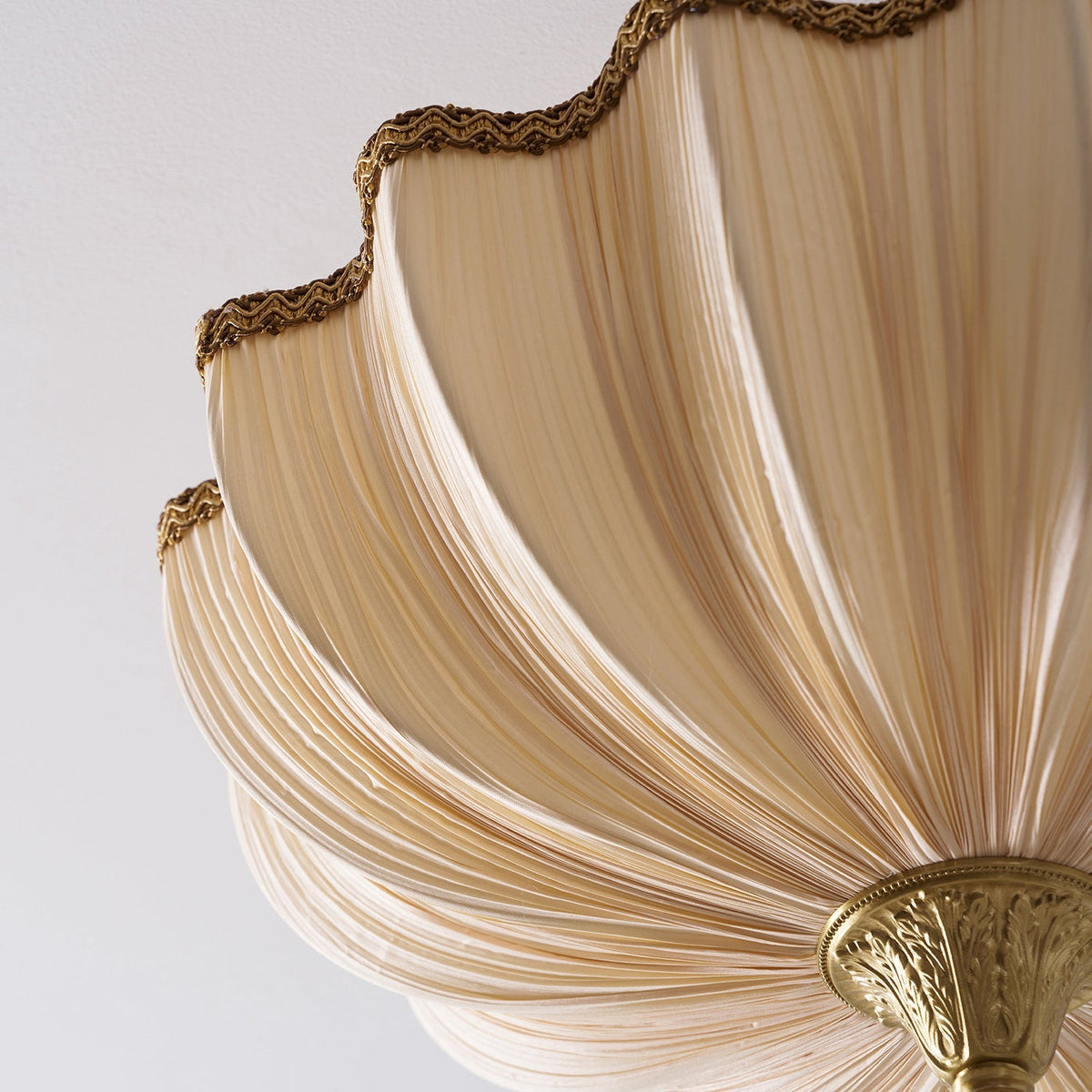 Close view of the braided scalloped trim and the pleated cream fabric on this Clérachic ceiling light, with the brass finial below.