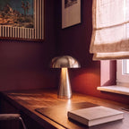 Clérachic silver table lamp glows on a sunlit desk, its dome shade warm beside a striped framed print.