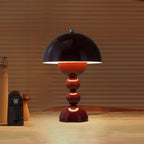 Wine red Clérachic dome lamp lit on a table, throwing a warm pool of light, with a wooden object and slatted panel behind.