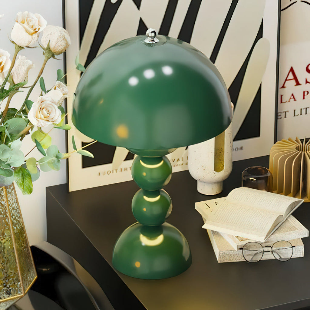 A green Clérachic dome lamp on a black desk, beside an open book, reading glasses, white roses and a graphic poster.