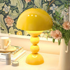 Yellow Clérachic dome lamp glowing on a pale desk, with a floral wallpaper, stacked books and a bunch of dahlias.