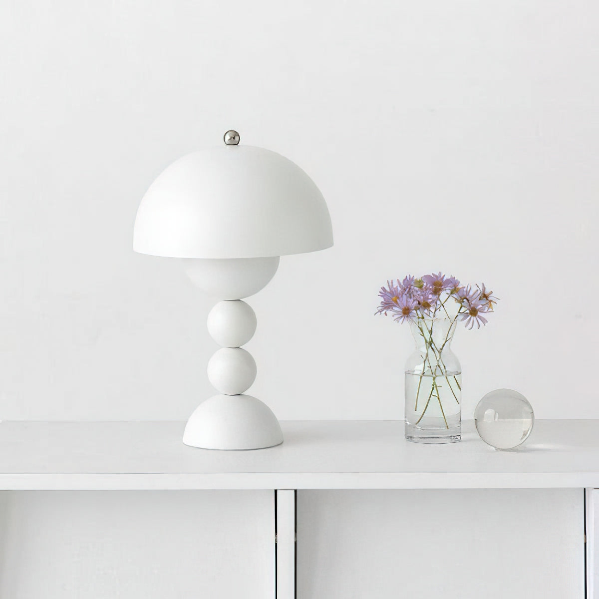 A white Clérachic dome lamp on a white console beside a small glass vase of purple flowers and a glass ball.