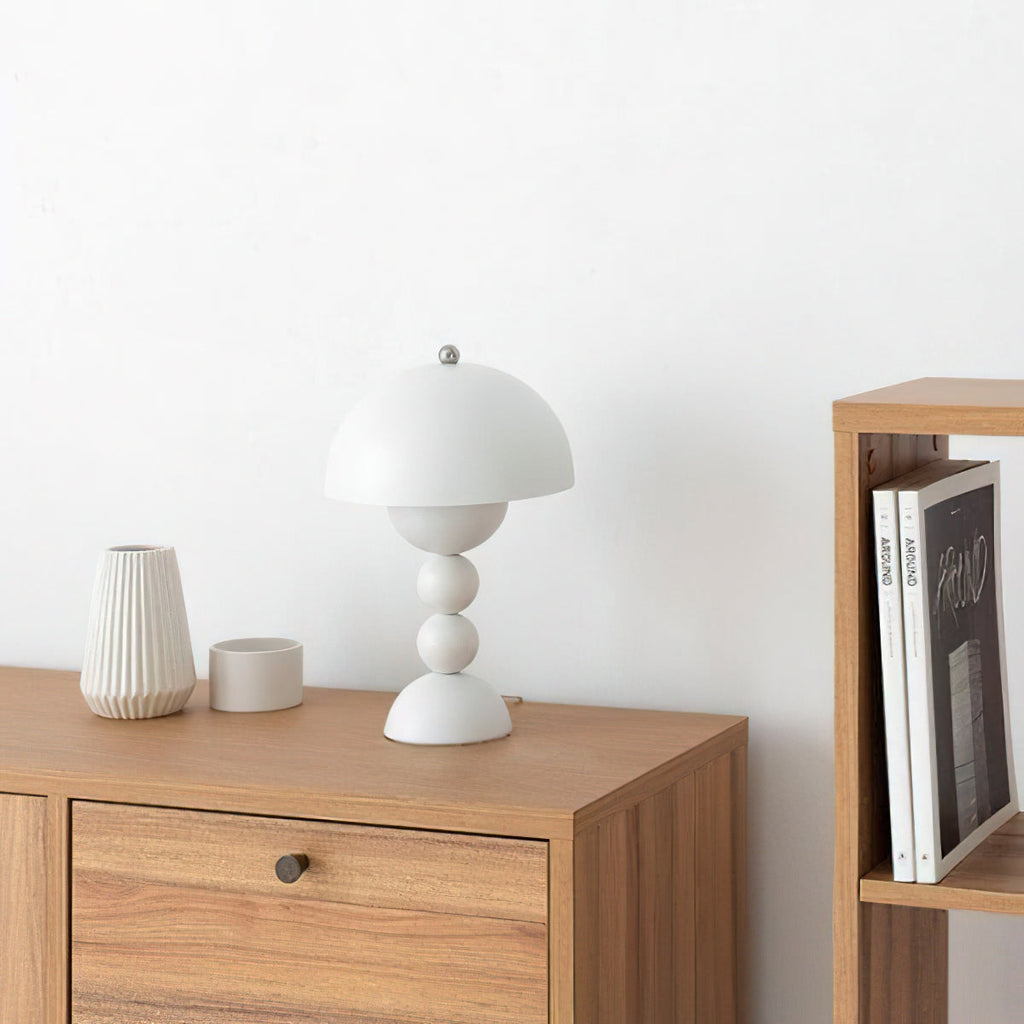 Small white Clérachic dome lamp on a wooden cabinet, with a ribbed vase, a small pot and books on a shelf nearby.