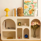 A small yellow Clérachic dome lamp sits on top of a cream arched shelving unit, with books, a basket and flowers below.