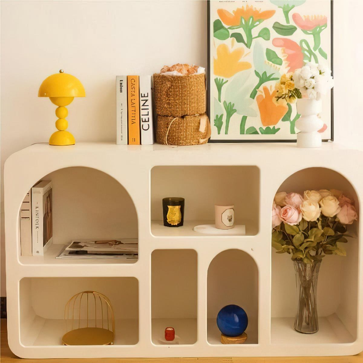 A small yellow Clérachic dome lamp sits on top of a cream arched shelving unit, with books, a basket and flowers below.