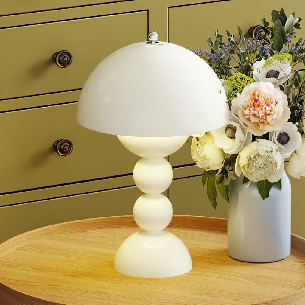 White Clérachic dome lamp lit on a round wooden table in front of olive drawers, beside a vase of peonies and anemones.