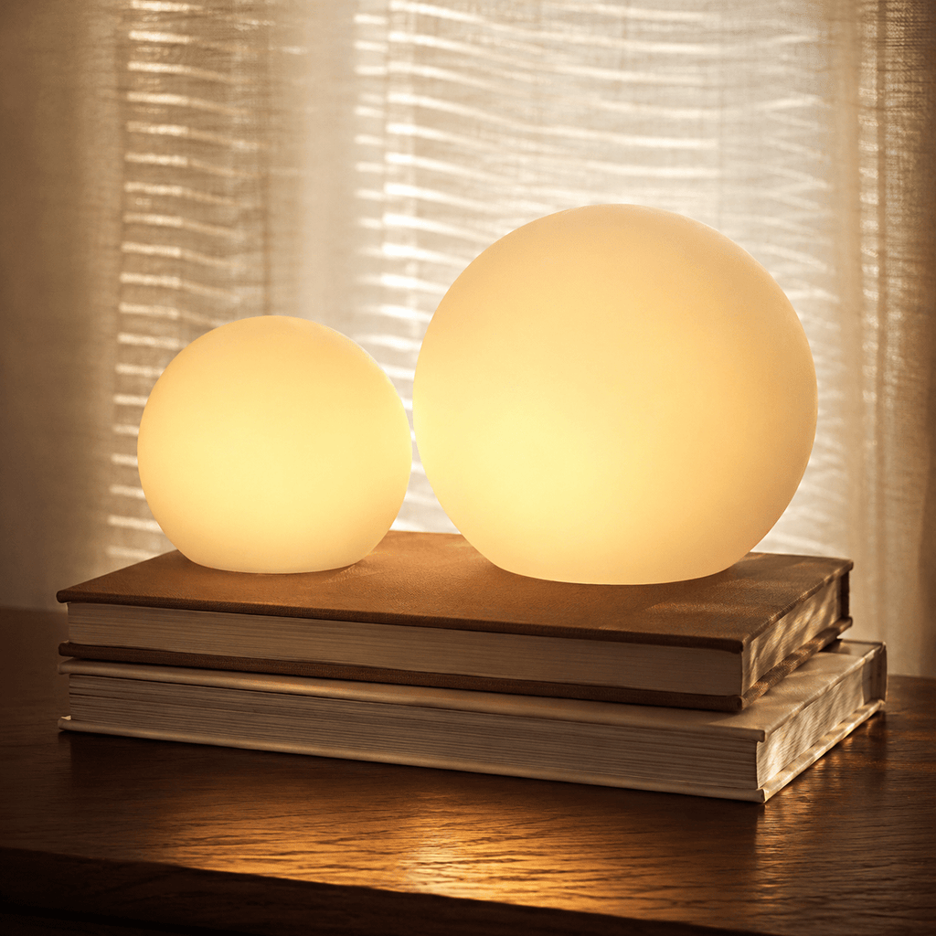 Two Clérachic orb lamps lit on stacked books, sheer curtains catching the light behind.