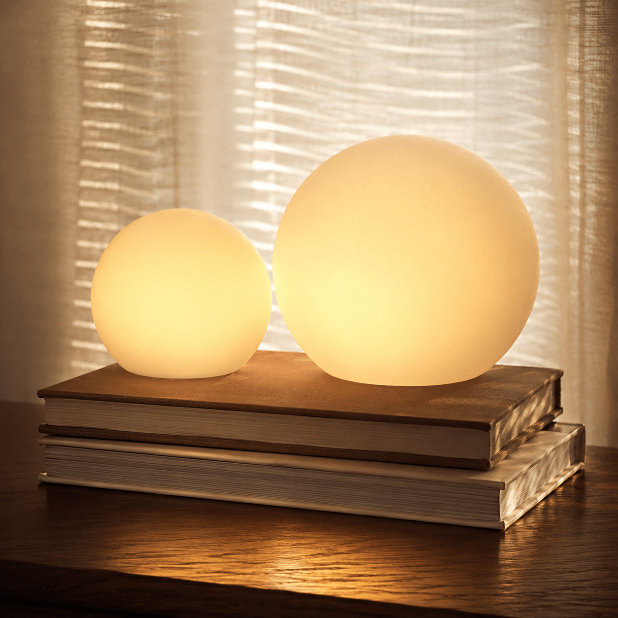 Two Clérachic orb lamps lit on stacked books, sheer curtains catching the light behind.