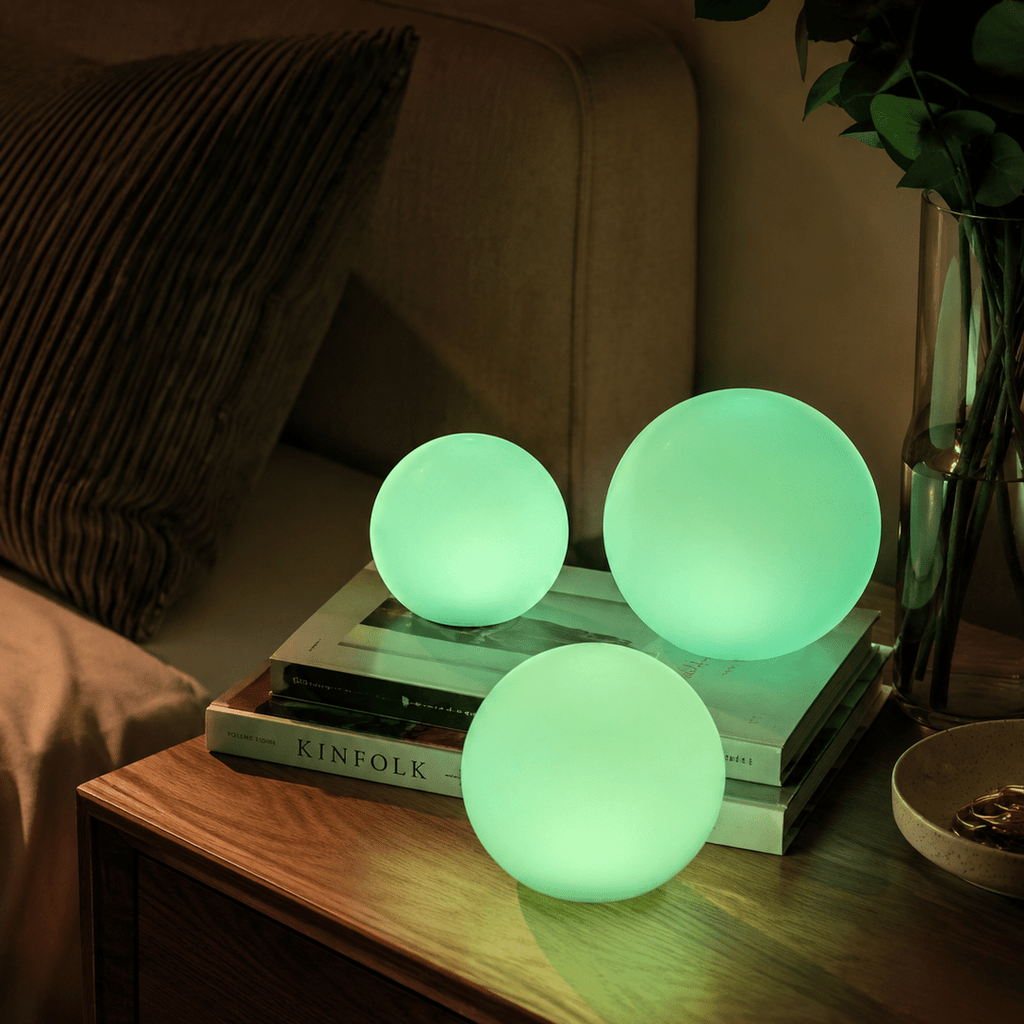 Three Clérachic orb lamps lit green on books and a bedside table with flowers behind.
