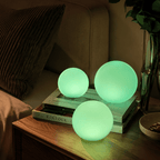 Three Clérachic orb lamps lit green on books and a bedside table with flowers behind.