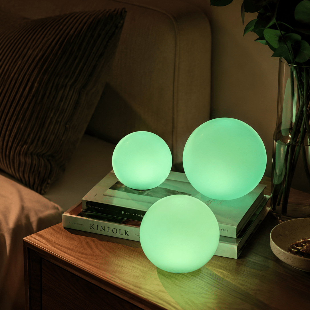 Three Clérachic orb lamps lit green on books and a bedside table with flowers behind.