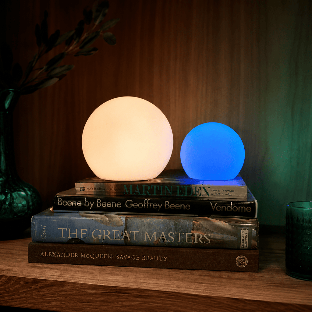 Clérachic orb lamps in warm white and blue, lit on a stack of art books against walnut.