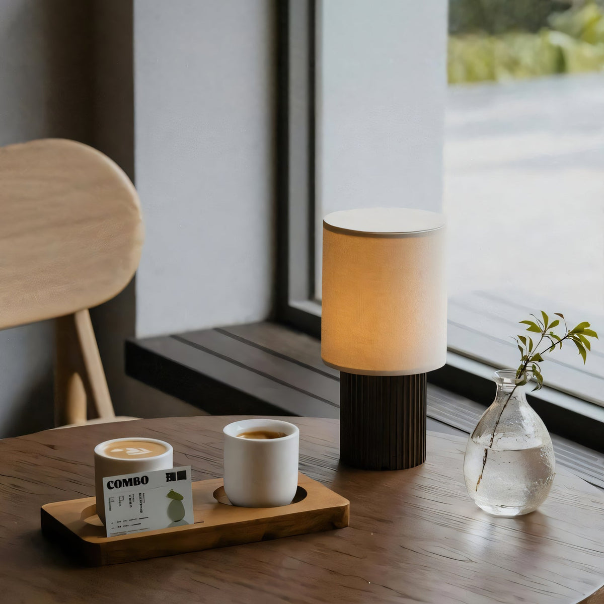 A Clérachic cordless lamp glowing on a dark wooden café table beside two coffee cups and a glass bud vase by a window.