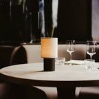 Dusk scene with this Clérachic table lamp lit on a round restaurant table, wine glasses and a curved banquette behind it.