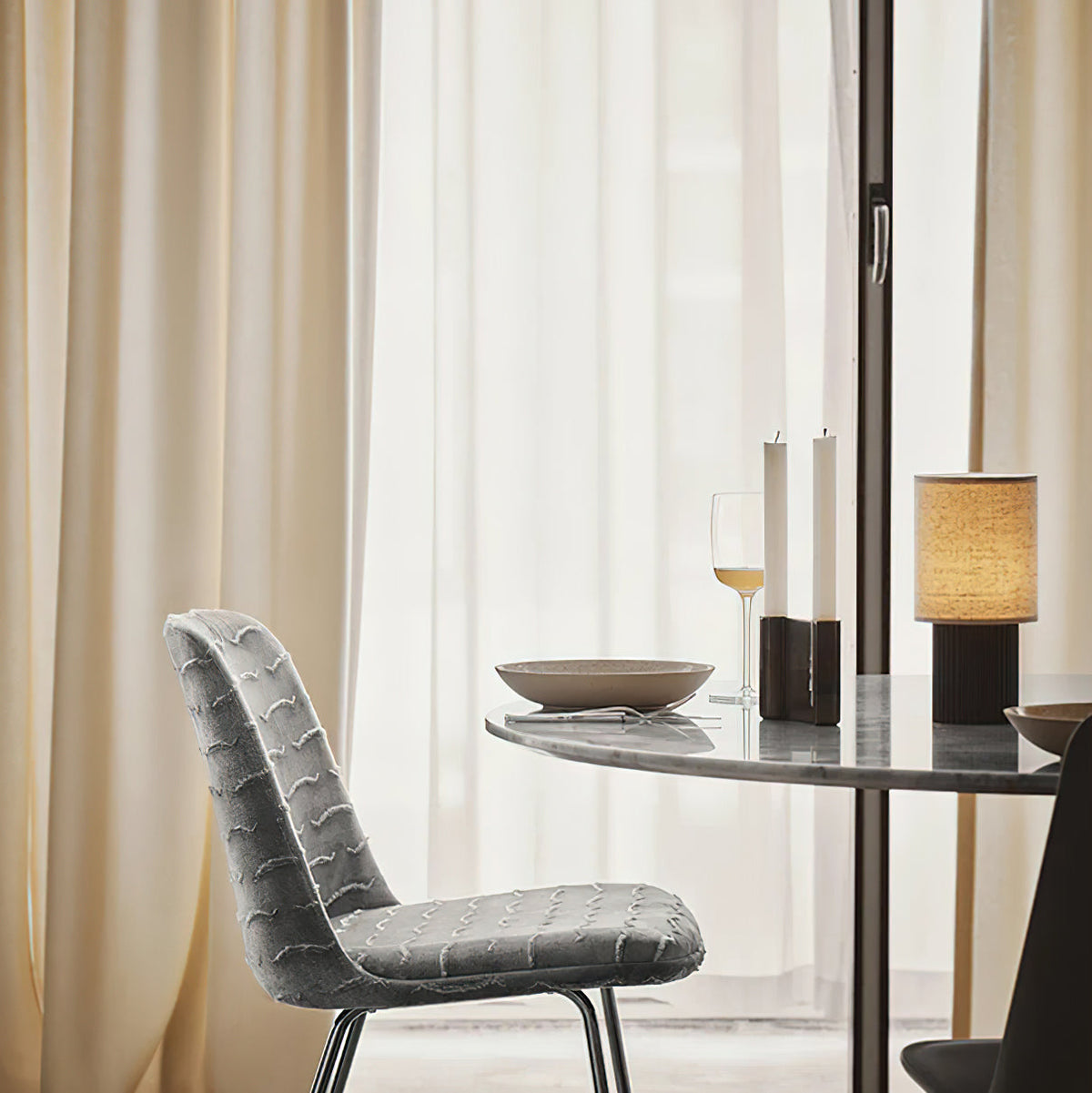 A Clérachic table lamp lit on a small round dining table by sheer curtains, with candles and a patterned grey chair nearby.
