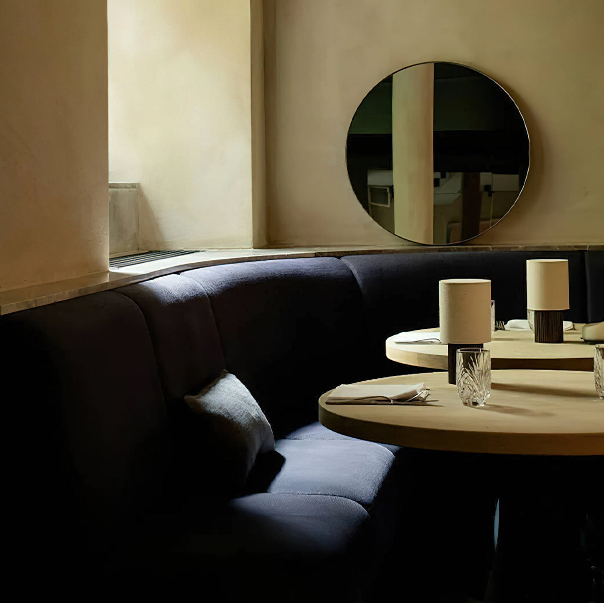 Small Clérachic lamps lit on café tables in the distance, with a round mirror and a dark curved sofa in the room.