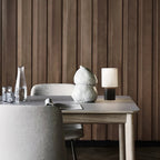 This Clérachic cordless lamp on a light wood table in front of vertical timber panelling, with a textured vase and a magazine.