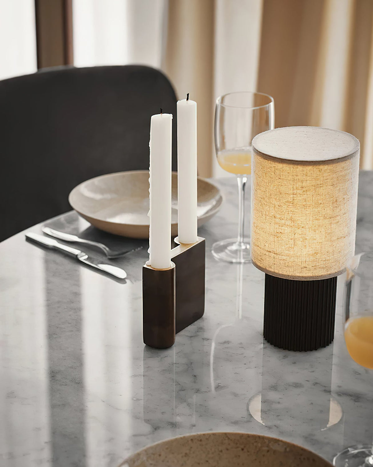 Linen shade of this Clérachic lamp glowing on a marble table beside tall white candles, a plate and a champagne glass.