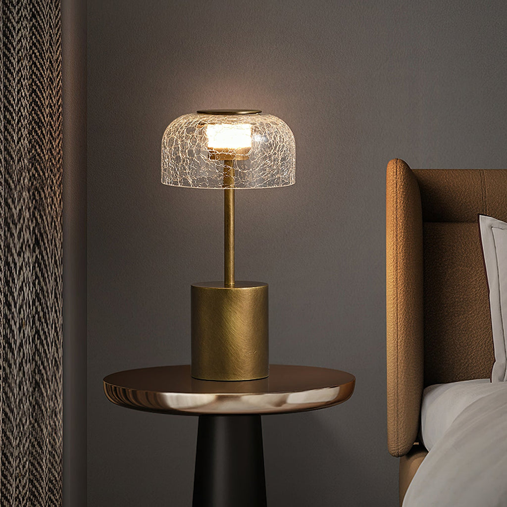 A lit Clérachic crackle glass table lamp on a small round side table beside a bed with a tan headboard, against a dark grey wall.